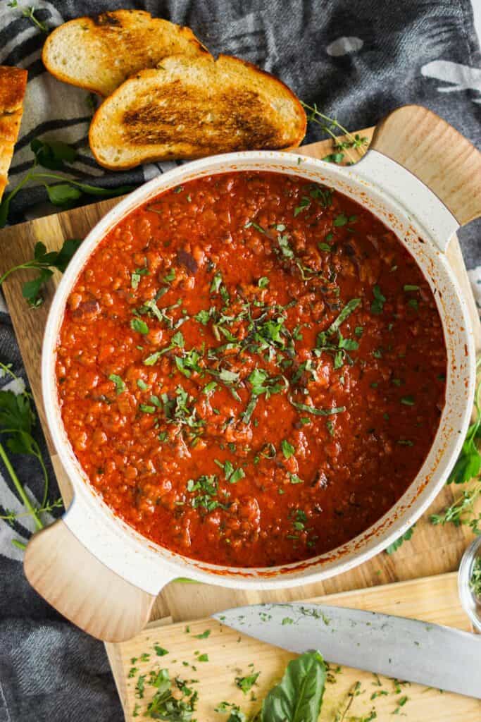 overhead view of a pot of bolognese sauce topped with parsley and basil, sitting on a cutting board with sliced toasted garlic bread.