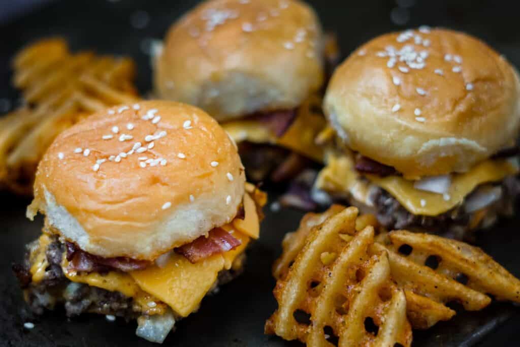 three bacon cheeseburger sliders on a dark pan with waffle fries