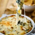 Spinach and artichoke dip in a white dish with a chip scooping some out with a cheese pull.