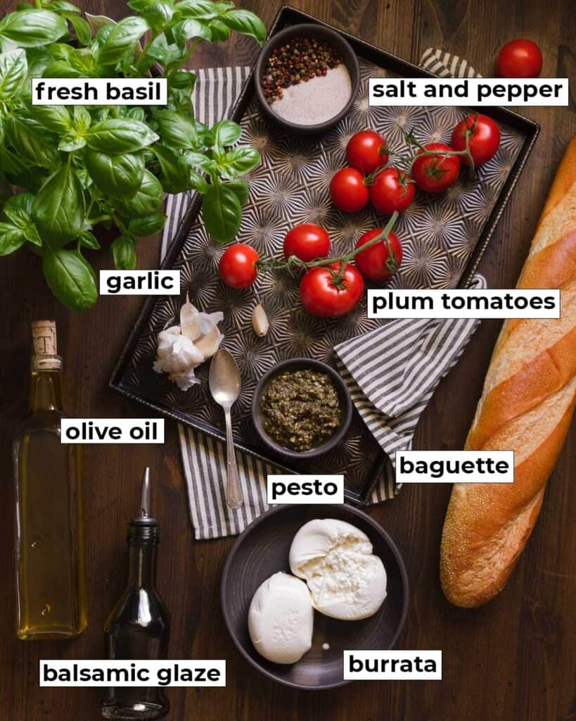 ingredients needed to make burrata bruschetta on a table, fresh basil, salt and pepper, garlic, plum tomatoes, olive oil, pesto, baguette, balsamic glaze, burrata