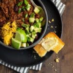 bowl of chili topped with avocado, cheddar, limes on a plate with corn bread