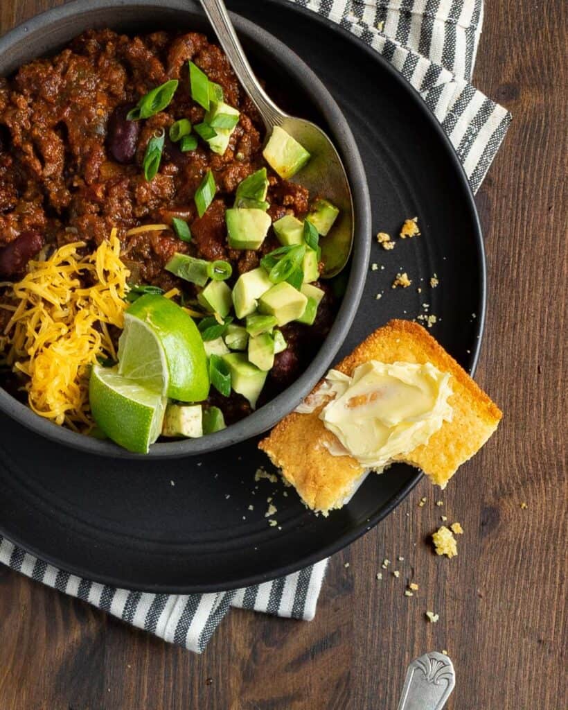 bowl of chili topped with avocado, cheddar, limes on a plate with corn bread