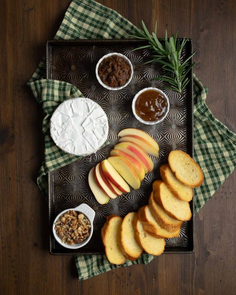 ingredients to make baked brie, fig jam, caramelized onions, apple slices, baguettes, rosemary, and walnuts on a tray