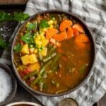 A close up shot of vegetable soup in a bowl with carrots, corn, parsley, green beans, potatoes, peas, and tomatoes.