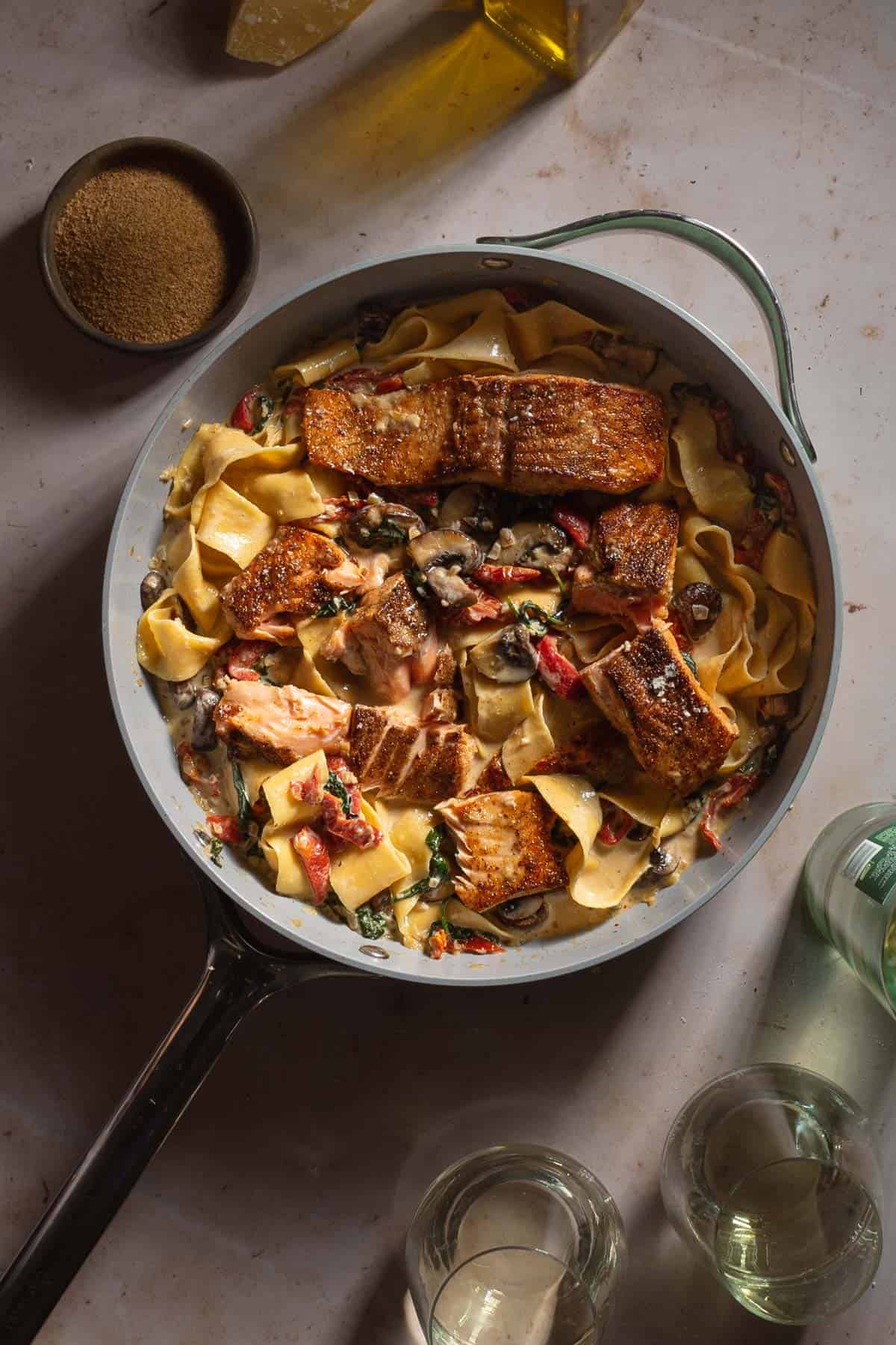 Cajun Salmon Alfredo Pasta in a skillet with pappardelle noodles next to wine glasses.