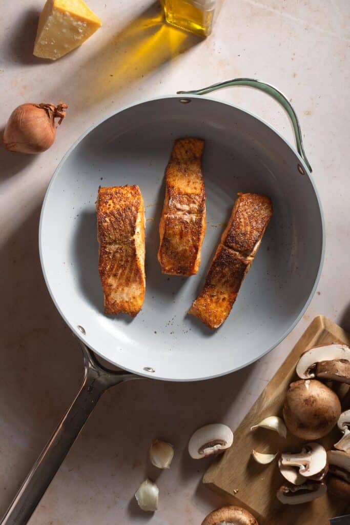 Seared Cajun salmon in a skillet next to a cutting board with mushrooms.