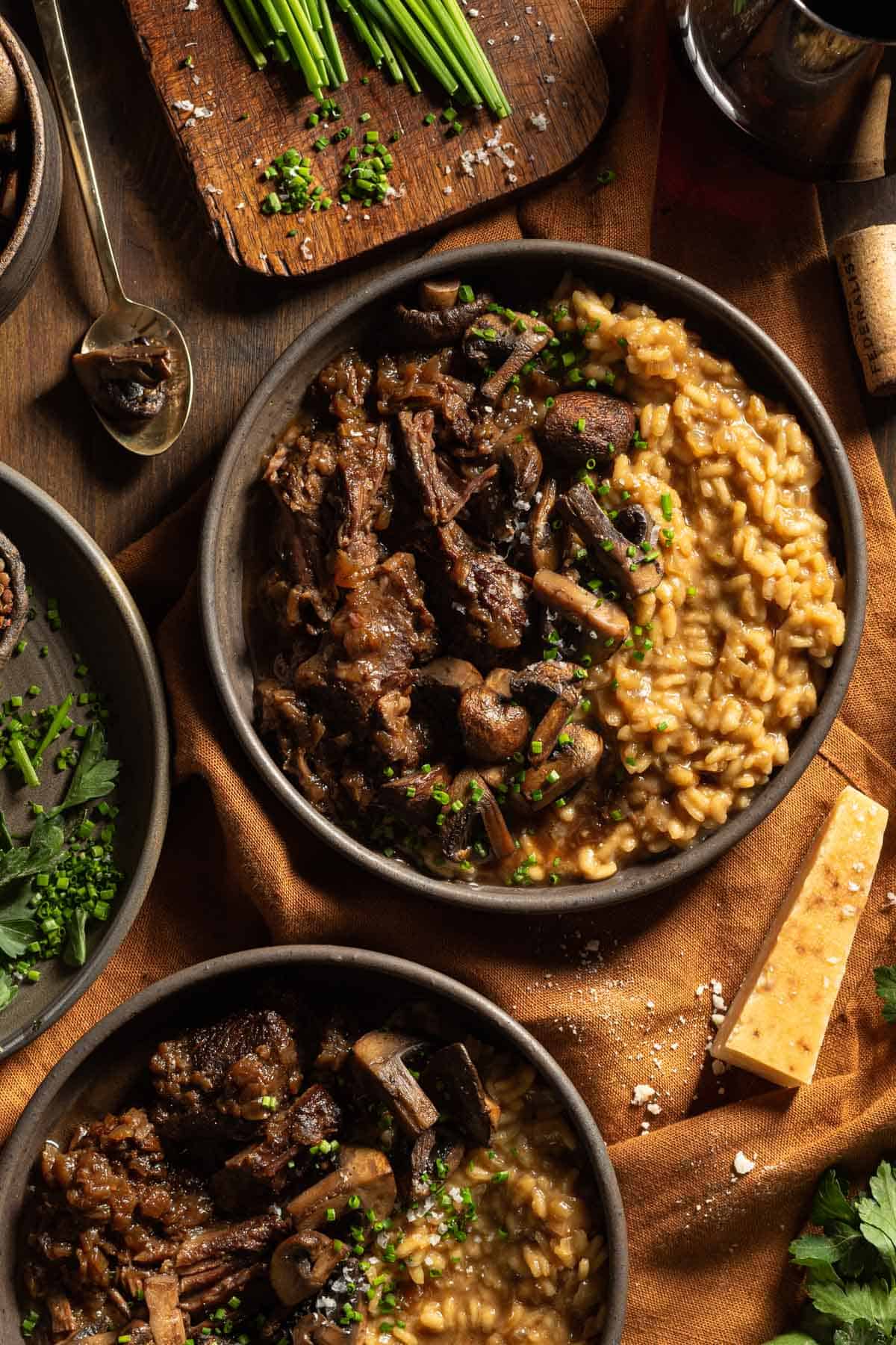 Two bowls of braised short ribs with mushrooms and risotto topped with chives on a table.