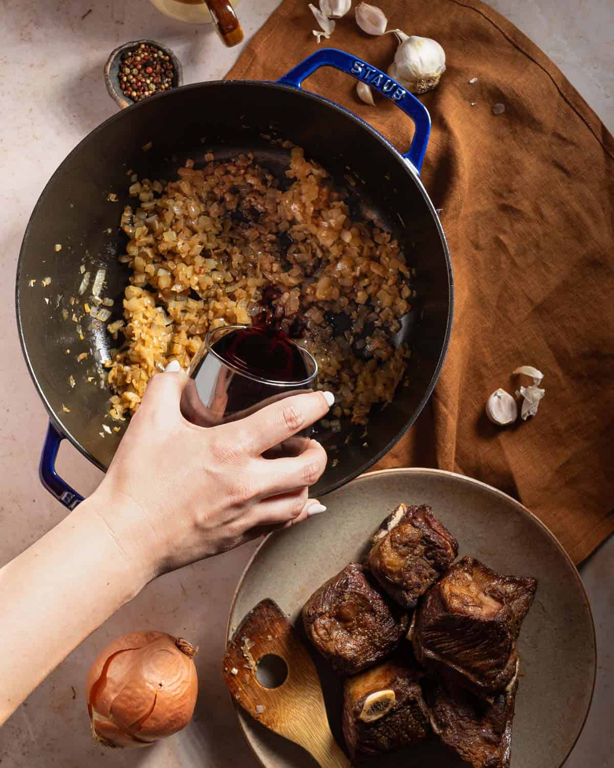 Adding red wine to deglaze a pot of sautéed onions next to a plate of seared short ribs.