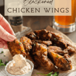 A hand dipping a chicken wing into ranch dressing with a plate of wings and a beer in the background.
