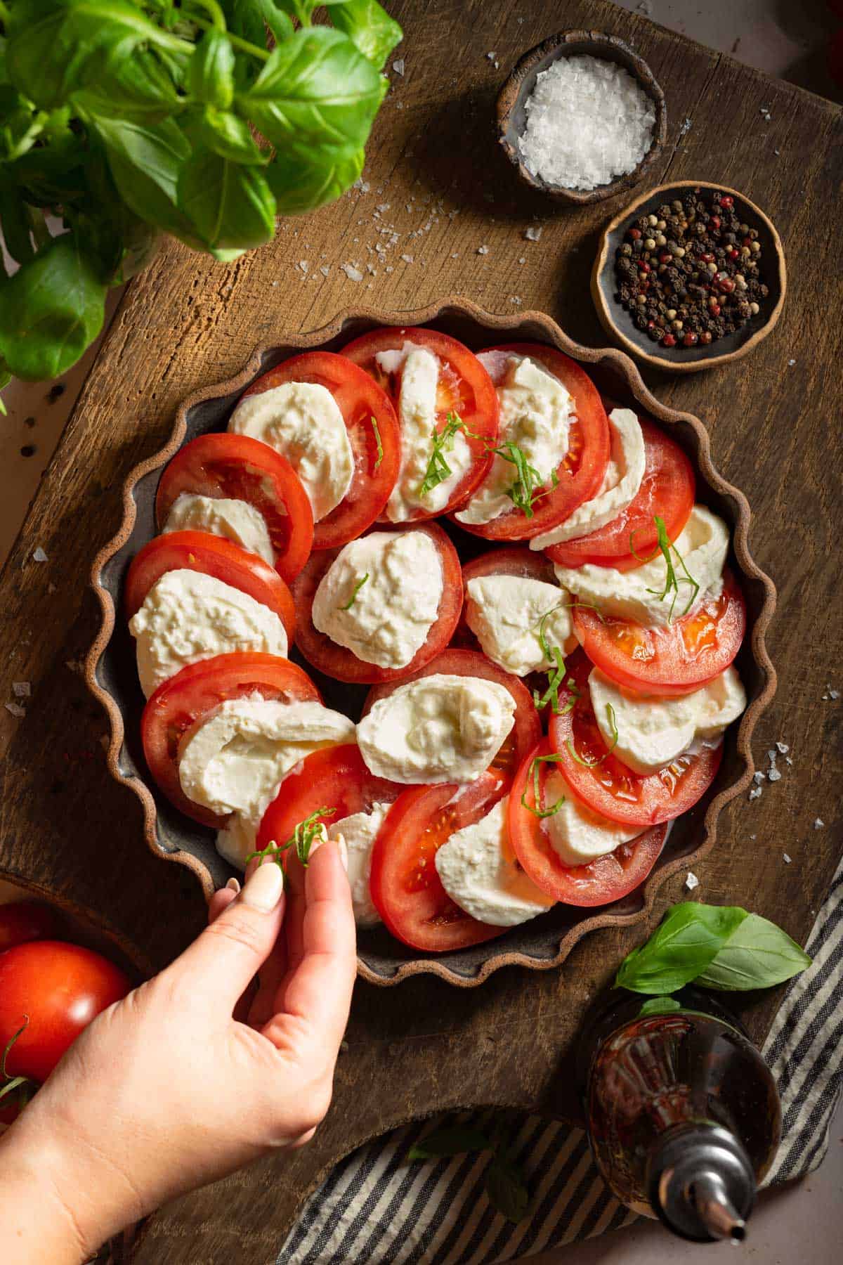 A hand adding fresh basil to a plate of burrata and tomatoes.