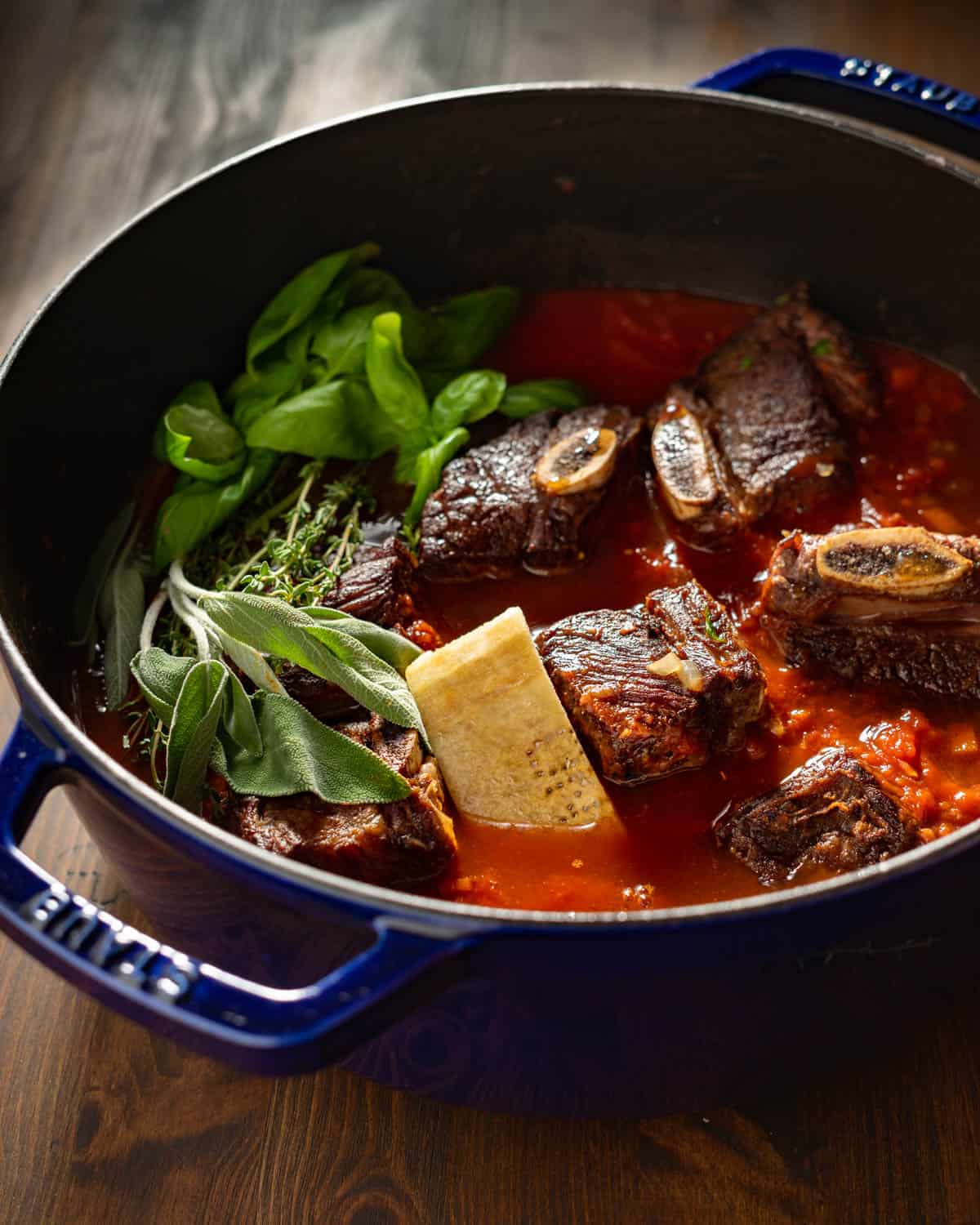 Short ribs with herbs, parmesan rind, and tomatoes in a large pot.
